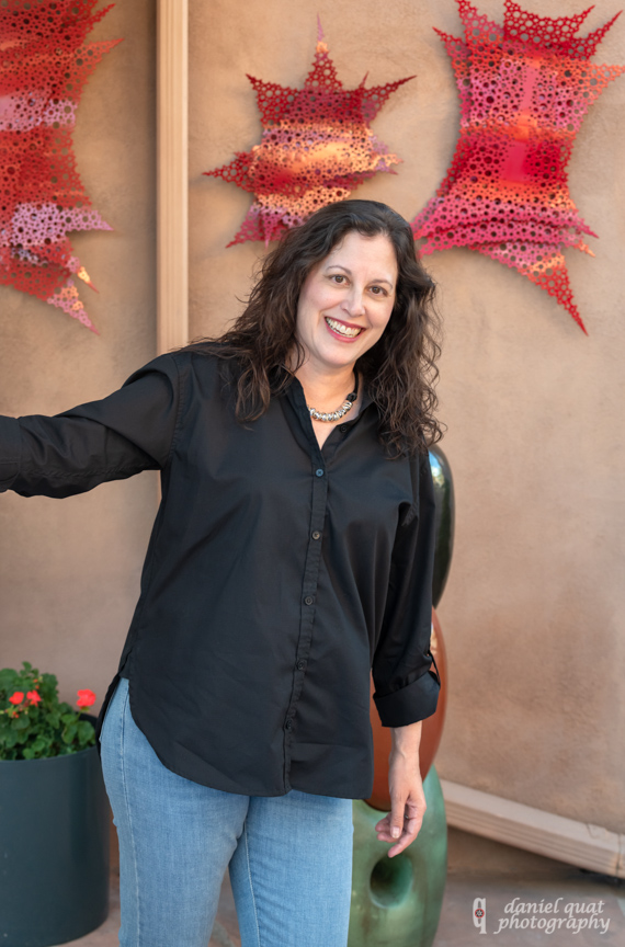 Michele Celeste Garcia photo by Daniel Quat taken in Santa Fe, New Mexico, shows Michele Celeste Garcia with long dark brown wavy hair and brown eyes wearing black long-sleeved blouse and silver necklace in front of orange and red artwork hanging on beige wall with orange geraniums in a pot on the ground behind her on the left side of the photo