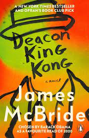 Off the shelf, Orange and yellow-green book cover with black line portrait of the outline of a man wearing a pork pie hat and suit jacket, with no facial features but instead the title of the book in black letters across the face area "Deacon King Kong," followed by "a novel" in small print with the name of the author, James McBride, in white letters across the chest area of the portrait. The words "Chosen by Barack Obama as a favorite read of 2020" are underneath the author's name, also in white letters.