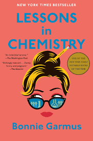 Off the shelf, Coral-colored book cover with sky blue lettering "Lessons in Chemistry" across the top in all caps with the author's name, Bonnie Garmus, in upper and lower case letters of the same color. In the middle of the cover is a drawing of a woman with blond-brown hair help up in a bun with a yellow pencil. She is wearing black semi-cat eye glasses with blue designs in them showing beakers and other chemistry equipment.