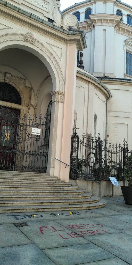 western entrance to the Sanctuary of the Consolata on Via Consolata in Turin on the morning of March 5, 2023, after the anarchists' protest the day and evening before. Painted in red are the words "Alberto libero" ("Free Alberto") and painted in black are the words "Porco Dio" ("God is a pig"). There are also black "paintball" stains on the walls of the church