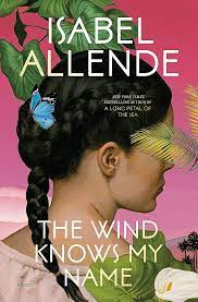 Book cover for The Wind Knows My Name by Isabel Allende, showing a young woman in profile from the back, with her eyes not visible, with a long, dark braid and a blue butterfly in her hair on a pink background