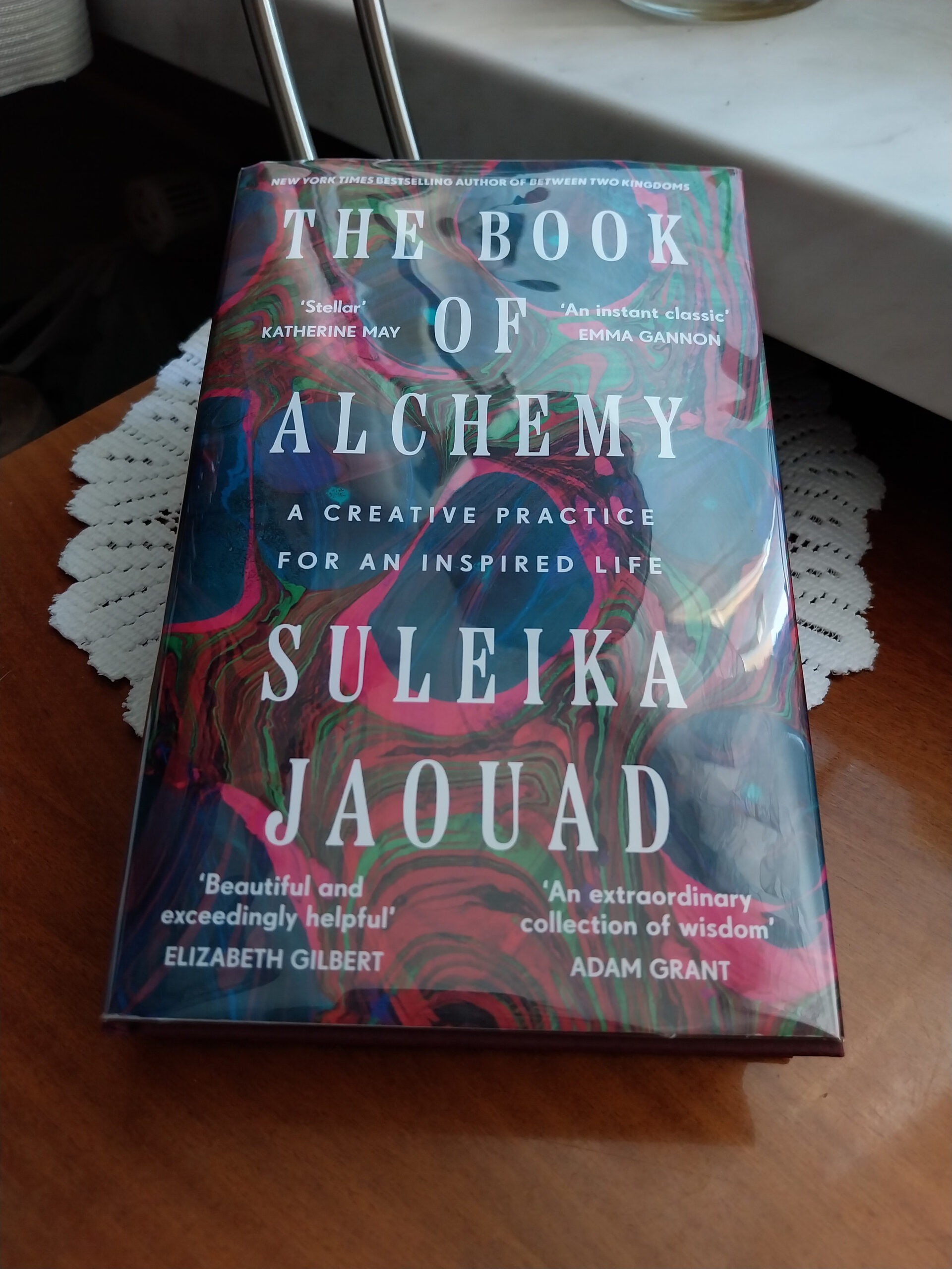 Creating Alchemy
Book cover for The Book of Alchemy: A Creative Practice for an Inspired Life by Suleika Jaouad. The cover shows the title and the author's name in large white font, with accolades by Katherine May, Emma Gannon, Elizabeth Gilbert, and Adam Grant printed onto the four corners of the cover. All of this is superimposed on a colorful background of medium blue cell-like structures with medium green, medium red, and hot pink lines swirled in between the blue shapes. The overall effect is one of blue blood cells surrounded by these multi-colored lines