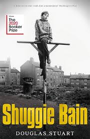Book cover for the 2020 Booker Prize winner Shuggie Bain, by Douglas Steward The cover is a black and white photograph of a young boy balanced on a kind of tall pole with a cross beam, his right leg hung over the horizontal bar and his left leg hanging down towards the ground. His hands are holding him upright and he's looking off into the distance. In the background are large apartment blocks with chimneys on a foggy morning. The look is desolate. In the near background is a trash can and the ground is devoid of grass or nature.